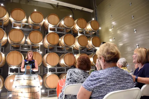 Presenter with wine barrels in the background and participants listening from the center aisle.